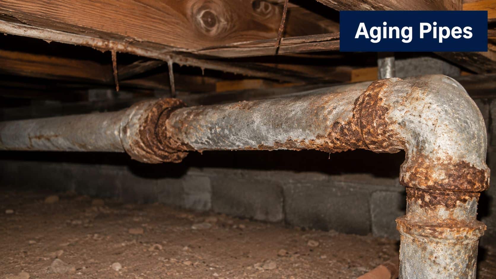 A close-up view of a corroded, rusted metal drain pipe beneath a house crawl space.